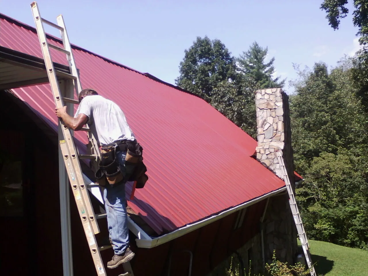 Roofer on a ladder beside a red metal roof and stone chimney — Soffit & Fascia Repair by Roofing Brothers in Wickliffe